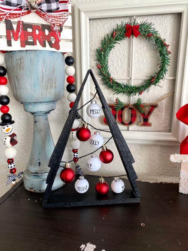 Black A frame wooden shim Christmas Tree with small red and white Rae dunn inspired bulb ornaments with a Christmas window wreath behind it and a snowman wood bead garland.