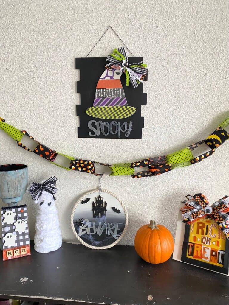 Halloween themed decorations on a bookcase with a witches hat, a haunted house beware sign, a mummy, a pumpkin, the trick or treat frame and a fabric banner.