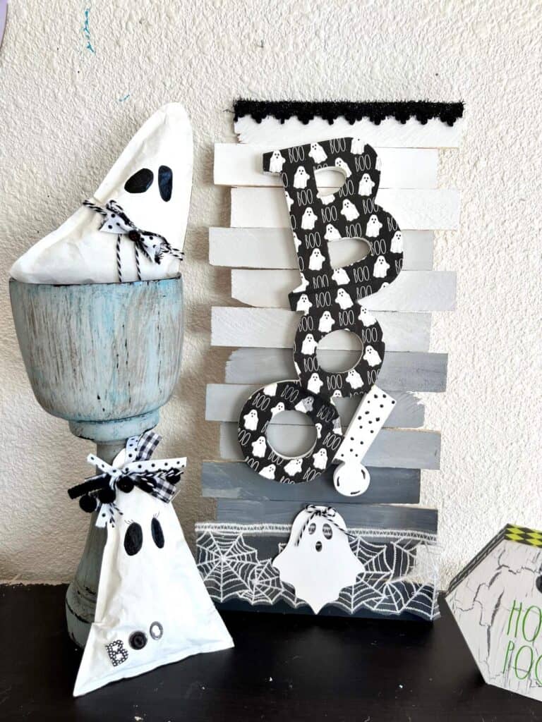 Dollar Tree BOO Sign made with a Rae Dunn Boo ghost napkin decoupaged and white, grey, black ombre background on shims with Halloween spiderweb trim. Sitting on a bookshelf next to kraft paper ghosts and a hocus pocus decor.