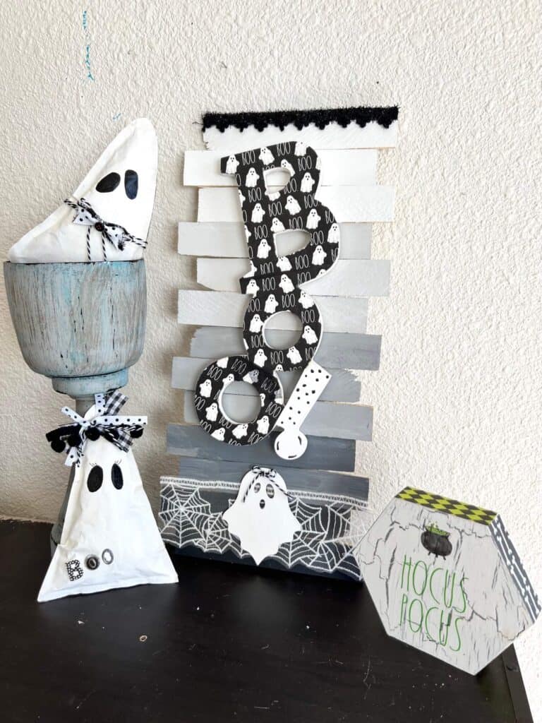 Dollar Tree BOO Sign made with a Rae Dunn Boo ghost napkin decoupaged and white, grey, black ombre background on shims with Halloween spiderweb trim. Sitting on a bookshelf next to kraft paper ghosts and a hocus pocus decor.