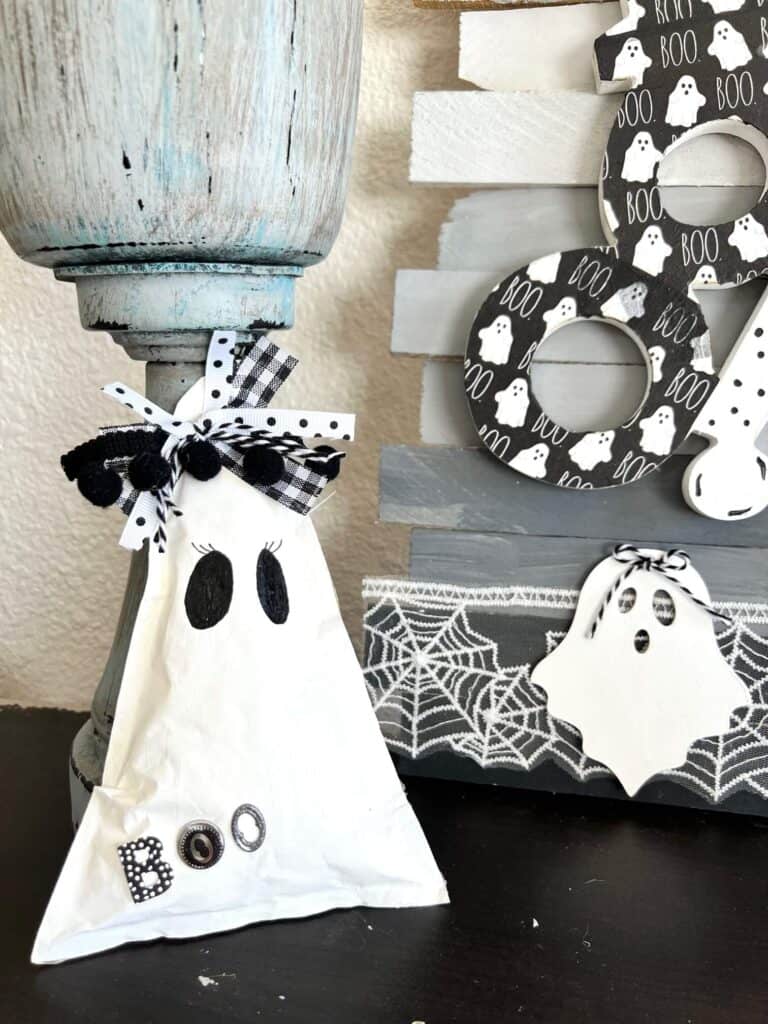 Girl Kraft paper ghost with a black and white bow on her head, black eyes with eyelashes, and the word "boo" with black, white and silver stickers, sitting next to a black white and grey BOO sign with small ghost.
