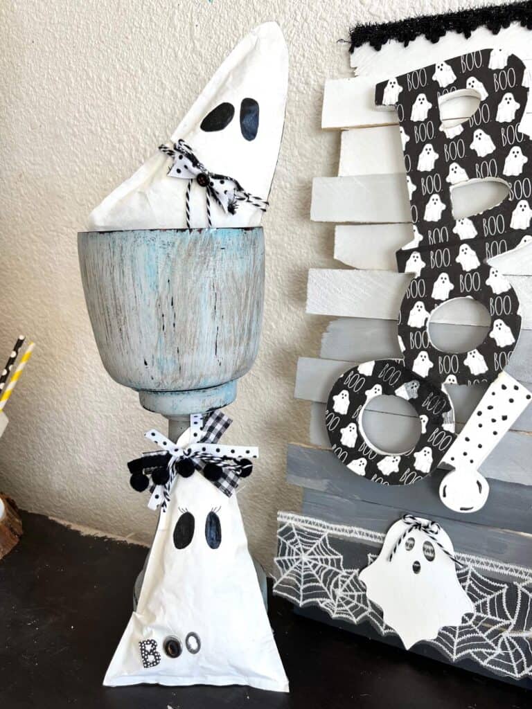 Dollar Tree BOO Sign made with a Rae Dunn Boo ghost napkin decoupaged and white, grey, black ombre background on shims with Halloween spiderweb trim. Sitting on a bookshelf next to kraft paper ghosts.