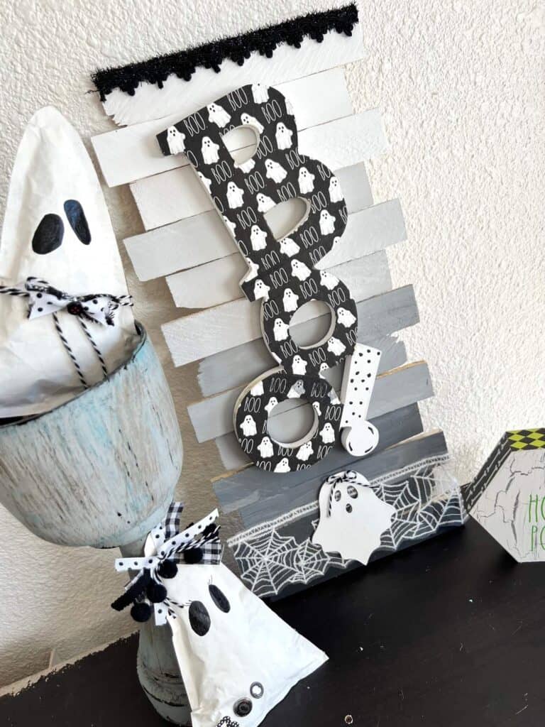 Dollar Tree BOO Sign made with a Rae Dunn Boo ghost napkin decoupaged and white, grey, black ombre background on shims with Halloween spiderweb trim. Sitting on a bookshelf next to kraft paper ghosts.