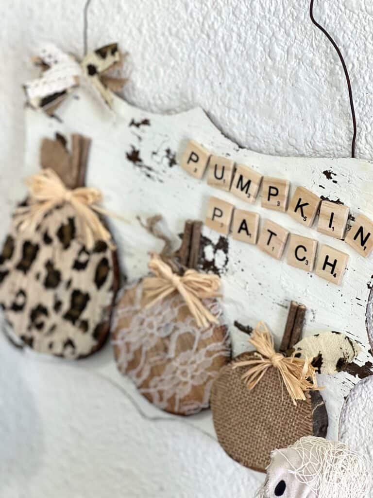 Rustic leopard and lace pumpkin patch made with 3 wood rounds and scrabble tiles and a wire hanger in a neutral theme with a white chippy paint background for fall decor.