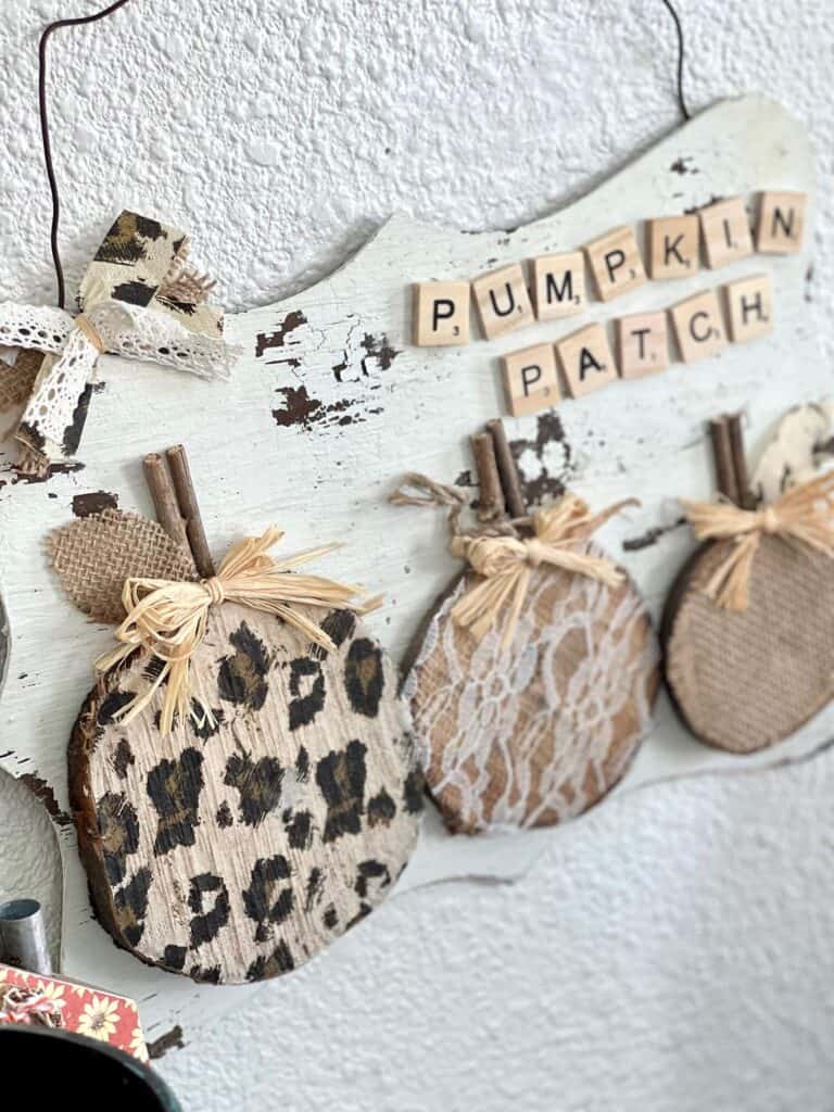 Rustic leopard and lace pumpkin patch made with 3 wood rounds and scrabble tiles and a wire hanger in a neutral theme with a white chippy paint background for fall decor.