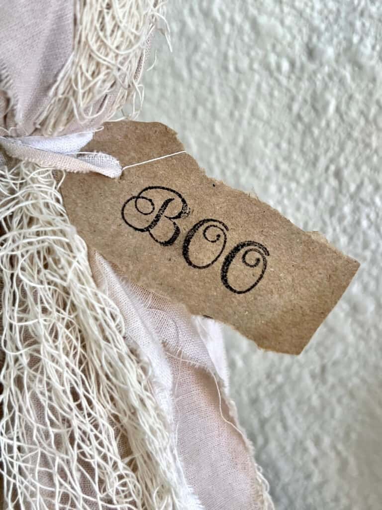 A ripped piece of Kraft paper tied around the ghosts neck with the word BOO in blank ink.
