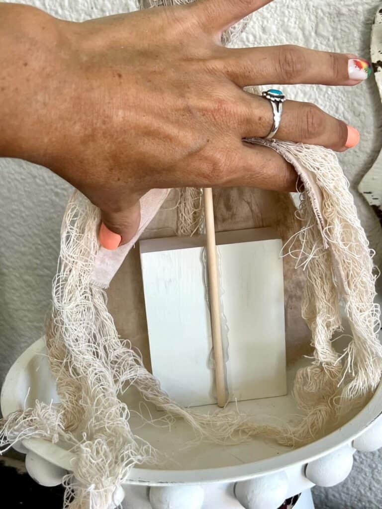 Hands lifting up the muslin and cheesecloth to see the wooden block that is the base of the ghost project with a wooden dowel glued to it.