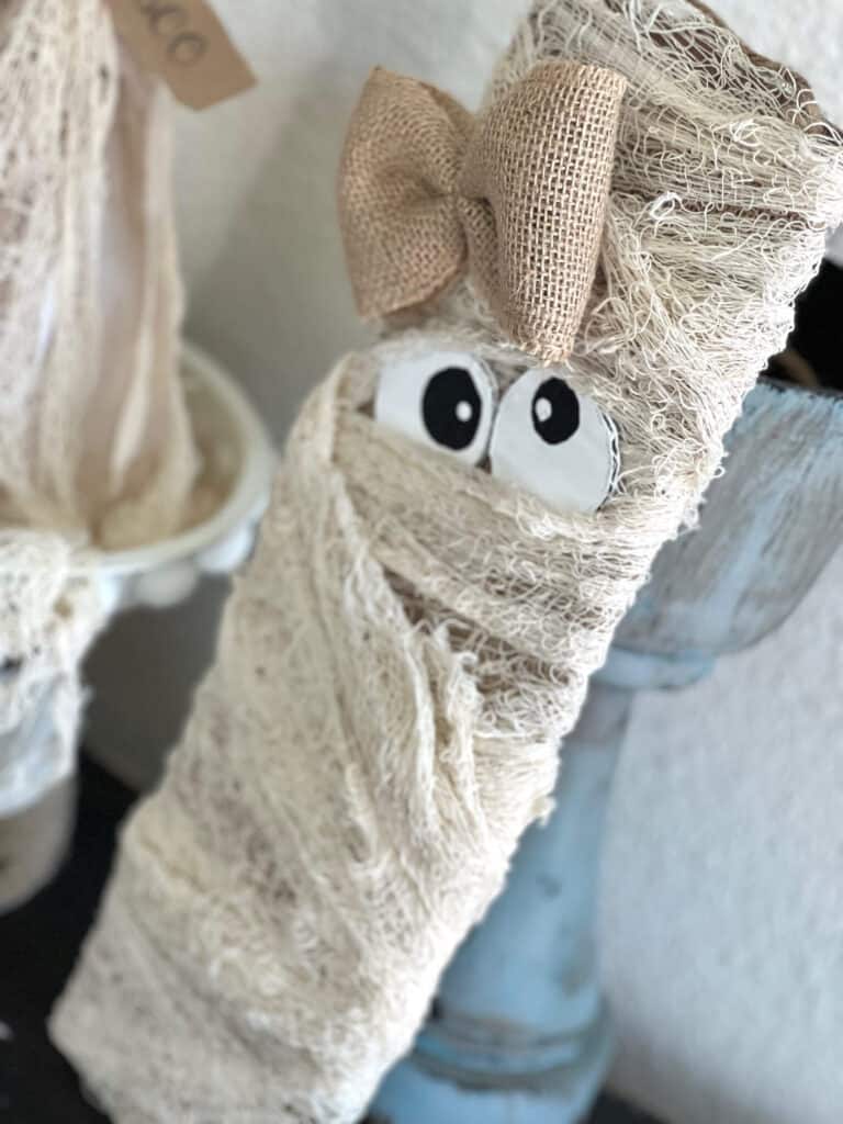 Dollar Tree Creepy Cheese Cloth Mummy Decor with burlap, a girly bow, cardboard wonky eyes on a long wood board for DIY Halloween decor on a budget.