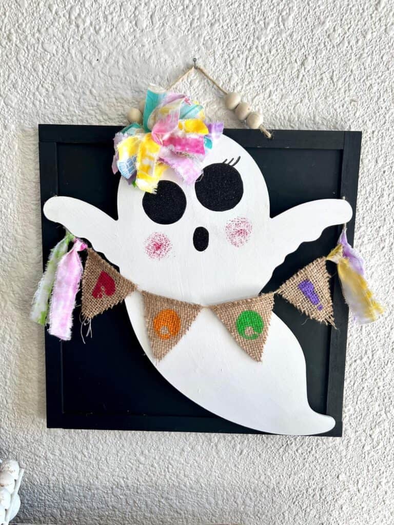 Dollar Tree Wooden Ghost with a Burlap Rainbow BOO Banner with colored fabric tassels, a rainbow messy bow, and big black sparkly ghost eyes with eyelashes, for cute DIY Halloween decor on a budget.