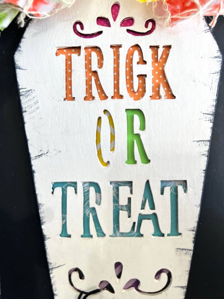 The words trick or treat cut out of the wood coffin with rainbow scrapbook paper behind it showing the color through.