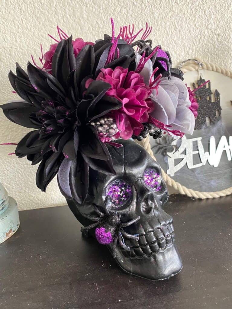 Dollar Tree Halloween Skull centerpiece with black skull, purple glitter eyes, a black and purple sparkly spider, and black, grey, and purple flowers for a spooky and glam piece of DIY Halloween decor for a home or a party.