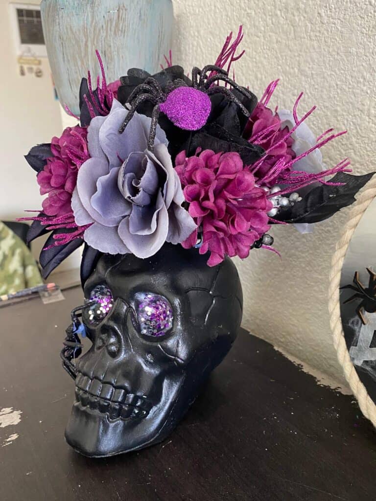 Dollar Tree Halloween Skull centerpiece with black skull, purple glitter eyes, a black and purple sparkly spider, and black, grey, and purple flowers for a spooky and glam piece of DIY Halloween decor for a home or a party.