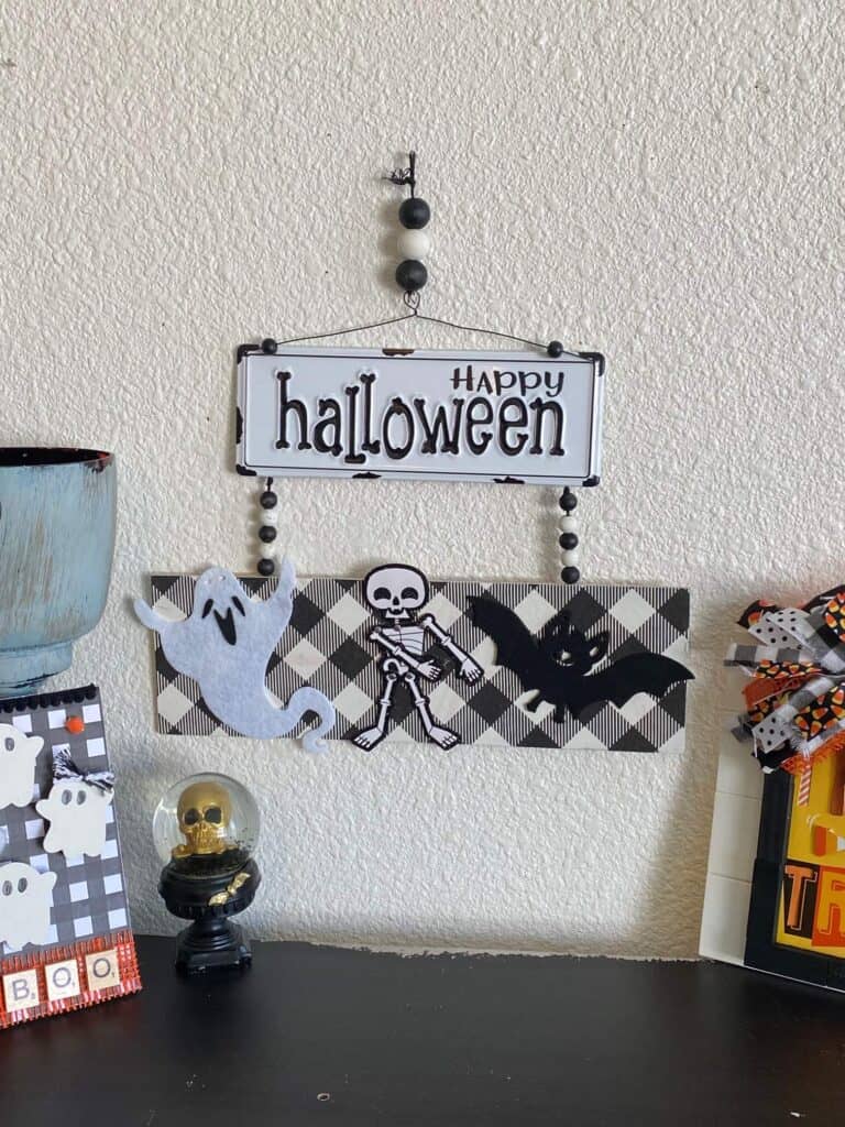 Happy Halloween Flossing skeleton DIY decoration with black and white metal sign and buffalo check along with a ghost, a bat, and a skeleton and wood bead hanger, hanging on the wall above a bookshelf with some other halloween decor.