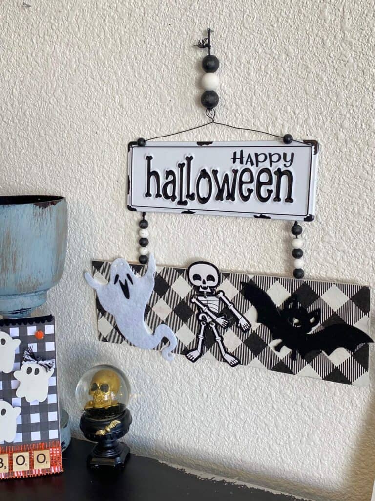 Happy Halloween Flossing skeleton DIY decoration with black and white metal sign and buffalo check along with a ghost, a bat, and a skeleton and wood bead hanger.