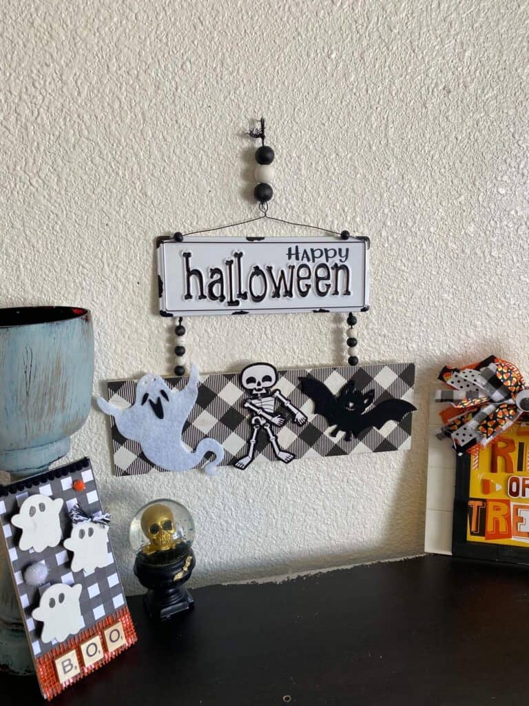 Happy Halloween Flossing skeleton DIY decoration with black and white metal sign and buffalo check along with a ghost, a bat, and a skeleton and wood bead hanger, hanging on the wall above a bookshelf with some other halloween decor.