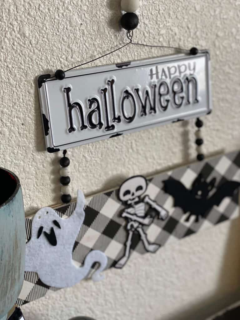 Metal Happy Halloween sign in black and white attached to a board with a ghost, skeleton, and a bat, with wood beads in between.