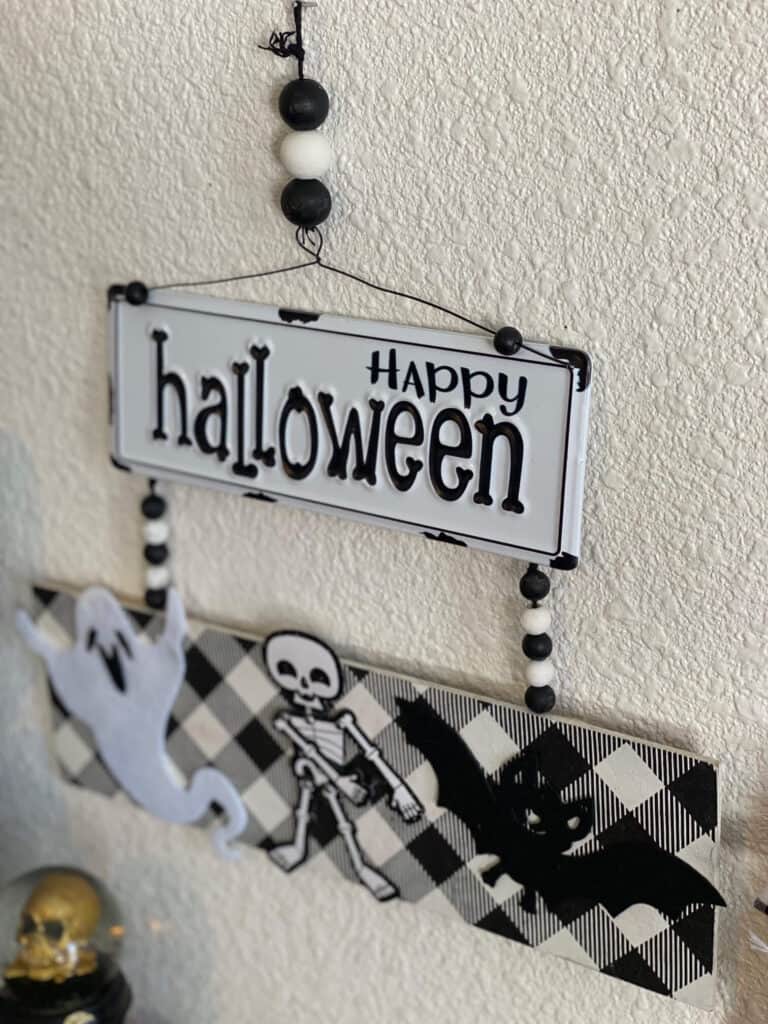 Metal Happy Halloween sign in black and white attached to a board with a ghost, skeleton, and a bat, with wood beads in between.