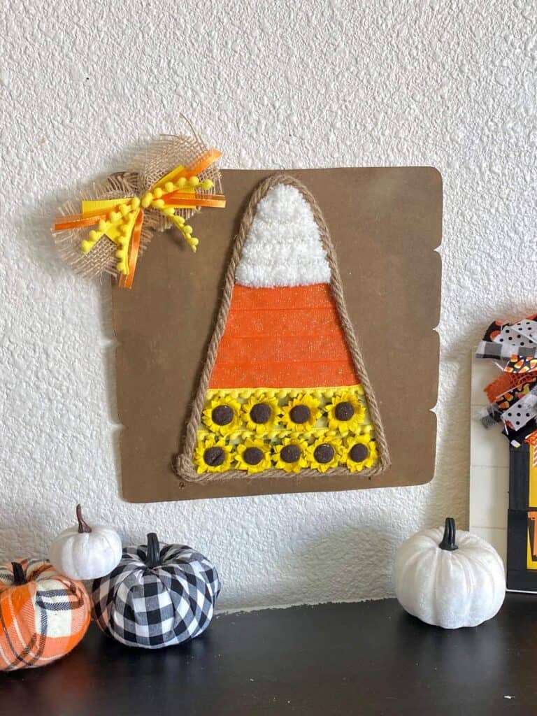 Dollar Tree Mixed Media Candy Corn craft with white chunky yarn, orange ribbon and yellow sunflowers with Jute rope around the edge and a cute bow in the corner. Mini pumpkins on the bookshelf underneath.