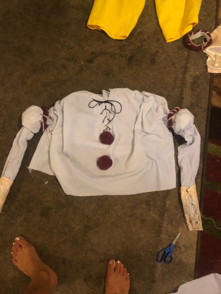 The white new pennywise shirt laying on the ground with large maroon pom poms glued to the front center and lace trim glued to the end of each sleeve.
