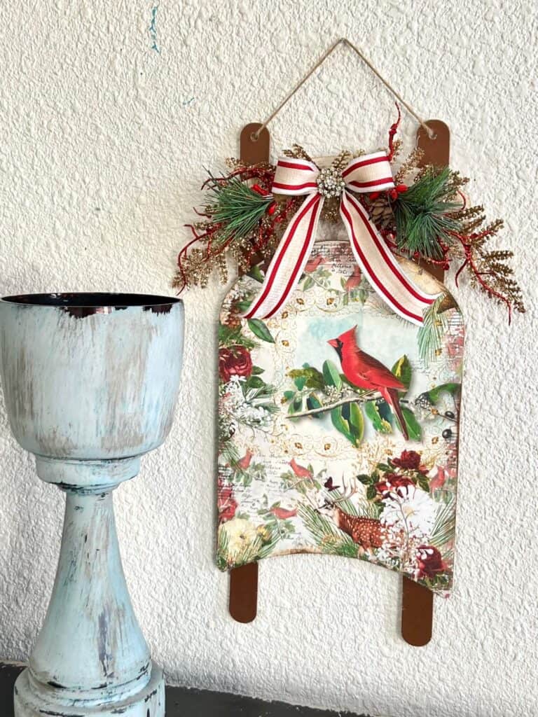 Dollar Tree Wooden Sled makeover for Christmas with a red cardinal rice paper print mod podged to the front, with greenery and a read and white striped bow and bling in the center for a Christmas Vintage DIY decor feel.