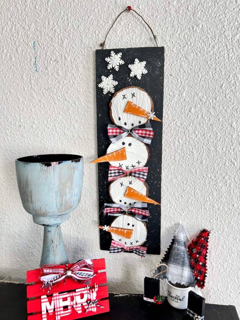 Wooden stacked snowmen decor made with wood round and 4 snowmen faces stacked on top of each other with bows in between on a black snowy background with a few snowflakes to decorate for winter.
