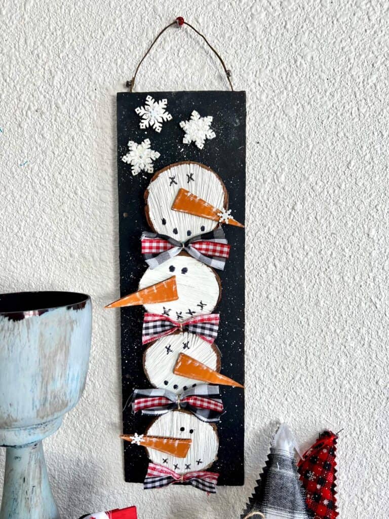 Wooden stacked snowmen decor made with wood round and 4 snowmen faces stacked on top of each other with bows in between on a black snowy background with a few snowflakes to decorate for winter.