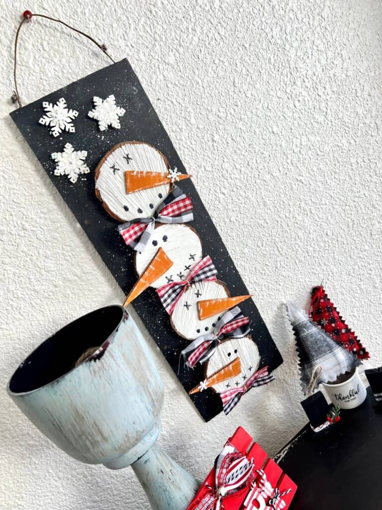 Wooden stacked snowmen decor made with wood round and 4 snowmen faces stacked on top of each other with bows in between on a black snowy background with a few snowflakes to decorate for winter.