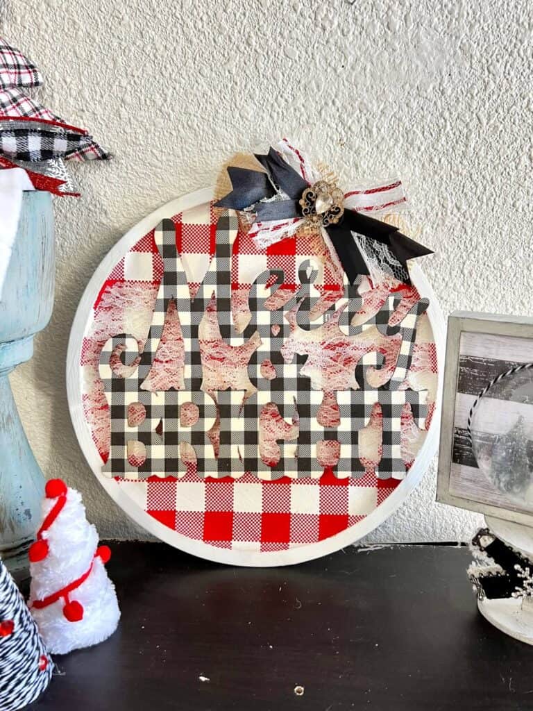 Dollar Tree Merry and Bright Christmas Pizza Pan Door Hanger Wreath with red and white check background and buffalo check wooden word cutout and a messy bow with vintage bling on a bookshelf next to some other DIY christmas decor.