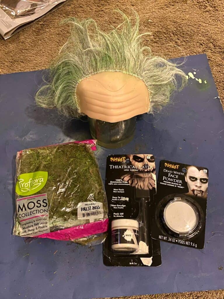 Half done Beetlejuice wig with green moss, Liquid Latex, and white powder on a blue poster board.