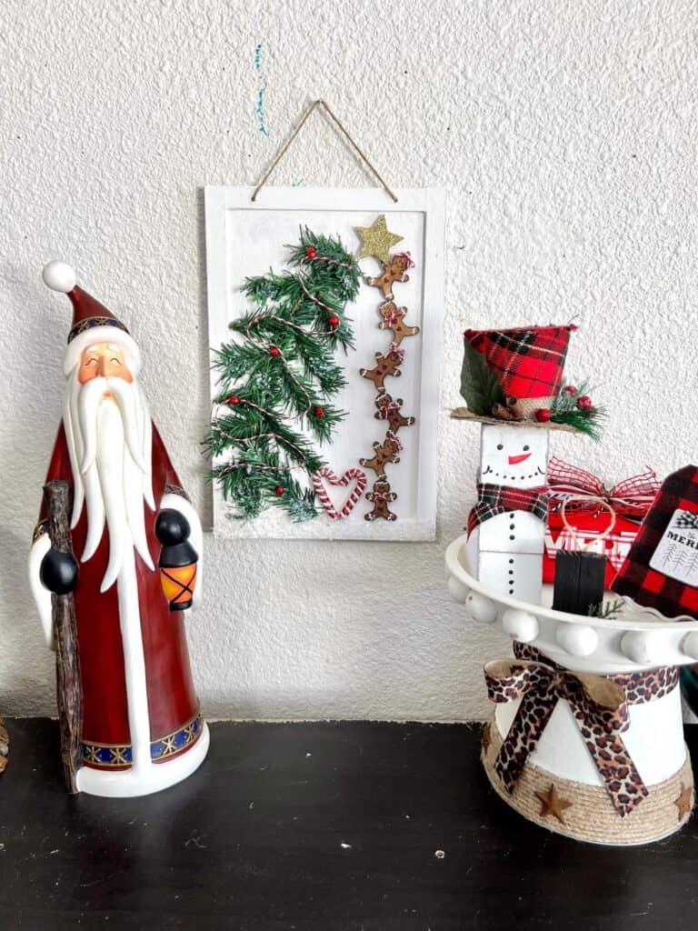 A tall standing Santa Claus figurine on a bookshelf next to the complete DIY Gingerbread Man craft in the middle with a small riser with a snowman on it to the right.