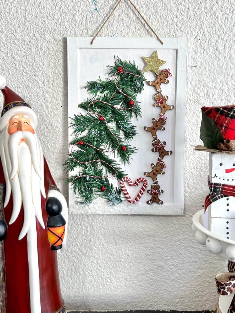 A tall standing Santa Claus figurine on a bookshelf next to the complete DIY Gingerbread Man craft in the middle with a small riser with a snowman on it to the right.