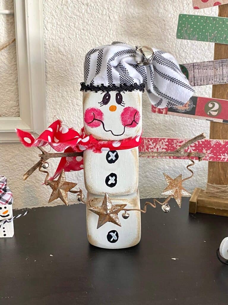 Dollar Tree Foam Dice Snowman DIY christmas and winter decor with a black and white striped hat, red polka dot scarf, sticks for arms, holding a jingle bell garland with rusty stars for decoration.
