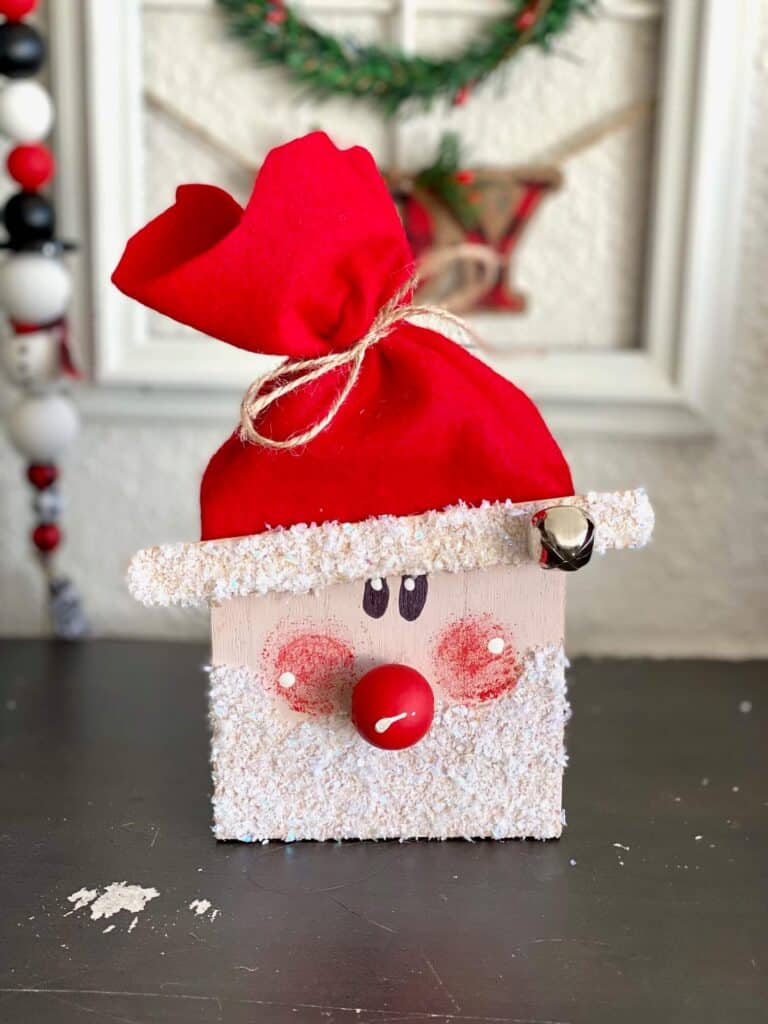 Wood block santa with faux glittery snow beard, red nose, red cheeks, and a red felt hat.