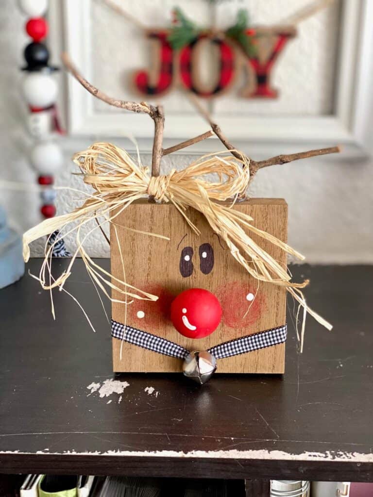 Wood block rudolph the red-nosed reindeer with stick antlers, a red wood bead nose, buffalo check collar with silver bell, and a raffia bow.
