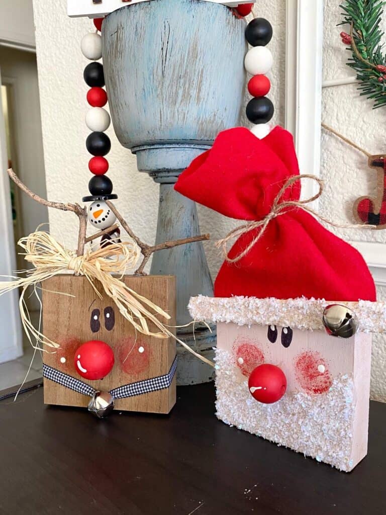 Wood block santa and rudolph the red-nosed reindeer made with 4x4 blocks, painted whimsy faces, wood bead red noses, raffia bow, and faux snowy glitter beard and jingle bells.