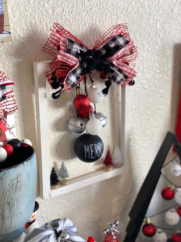 Hanging Christmas ornament reverse canvas made with red, black, white, and silver Rae Dunn inspired bulbs, hanging from black twine from a white 8x10 canvas with a big messy bow and mini bottle brush trees on the bottom.