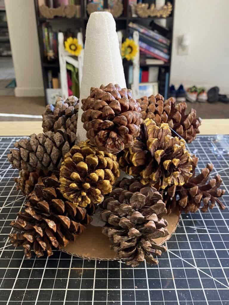 A foam cone glued to a circle cardboard base with pine cones glued all around the cone from the bottom up to about halfway up to the cone.