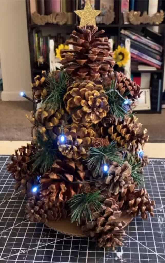 The completed pine cone christmas Tree with a string of white fairy lights and a gold glitter star on top.