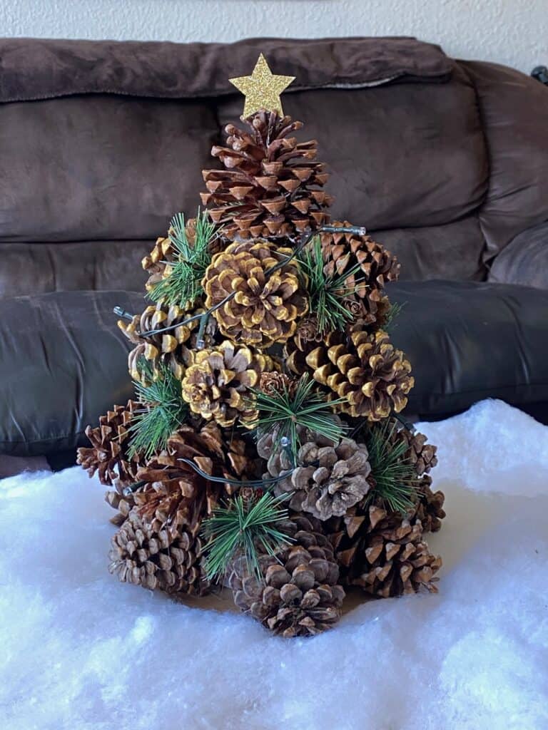 Pine Cone Christmas Tree with faux snow underneath, greenery added in, a string of white fairy lights, and a gold glitter star on top.