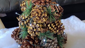 Pine Cone Christmas Tree with faux snow underneath, greenery added in, a string of white fairy lights, and a gold glitter star on top.