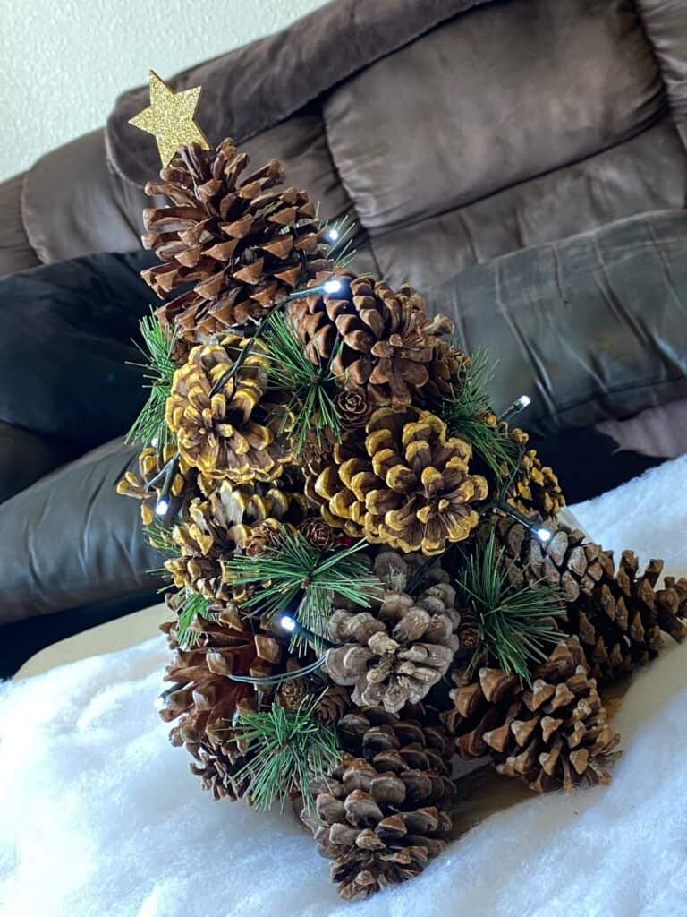 Pine Cone Christmas Tree with faux snow underneath, greenery added in, a string of white fairy lights, and a gold glitter star on top.