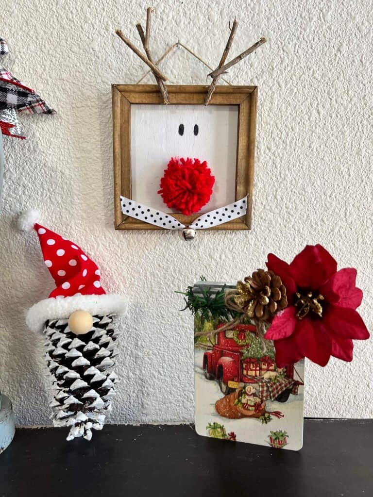 The staged craft project hanging on the wall with a pine cone gnome and a mason jar poinsettia on a bookcase.