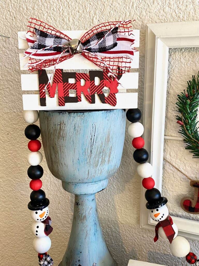 White pallet board with a red and black buffalo check Merry cutout from Dollar Tree with a coordinating bow and a silver bell on top of a chunky candlestick and a snowman wood bead garland.