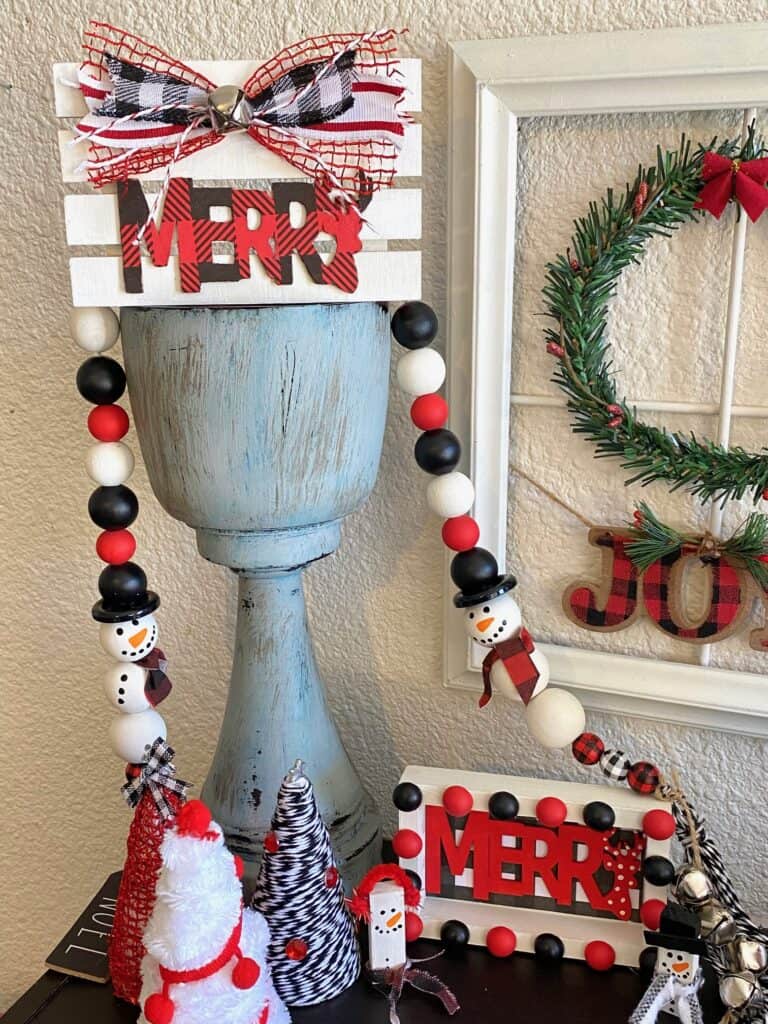 White pallet board with a red and black buffalo check Merry cutout from Dollar Tree with a coordinating bow and a silver bell on top of a chunky candlestick and a snowman wood bead garland.