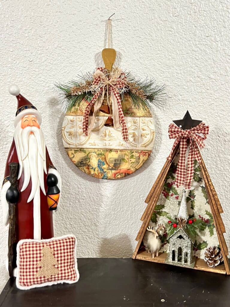 Tall santa ceramic statue, vintage ornament, and Wooden shim christmas tree all staged together on a bookcase and wall.