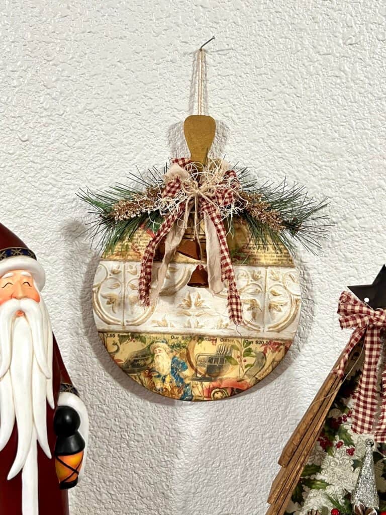 Vintage Christmas Bulb Ornament made with wooden cutout, Dollar Tree wall tiles, classic vintage g=holiday rice paper printable, a fabric bow with red homespun fabric, and some faux greenery and rusted bells.