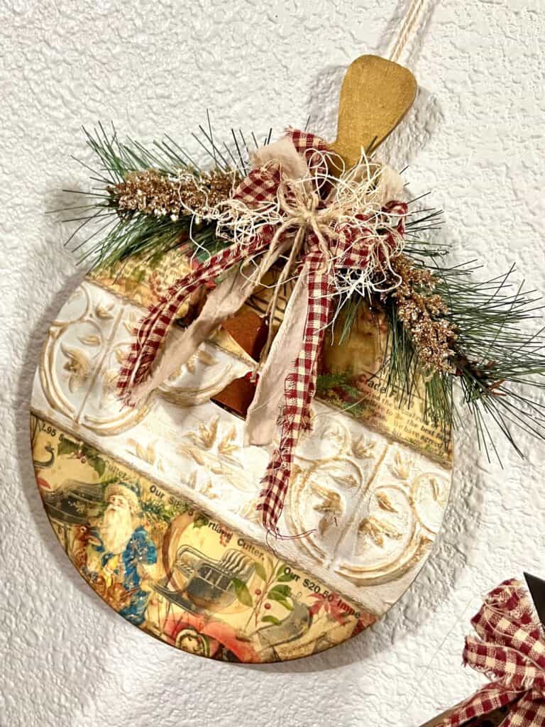 Vintage Christmas Bulb Ornament made with wooden cutout, Dollar Tree wall tiles, classic vintage holiday rice paper printable, a fabric bow with red homespun fabric, and some faux greenery and rusted bells.