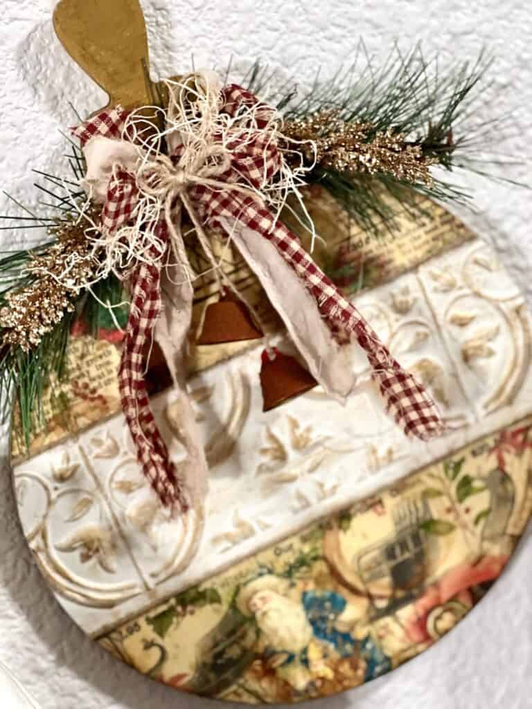 Vintage Christmas Bulb Ornament made with wooden cutout, Dollar Tree wall tiles, classic vintage holiday rice paper printable, a fabric bow with red homespun fabric, and some faux greenery and rusted bells.