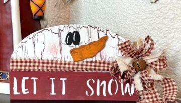 Dollar Tree half wood snowman with white paint and crackle paint underneath, cardboard carrot nose, homespun fabric scarf and bow, and the bottom portion is dark red and say let it snow.