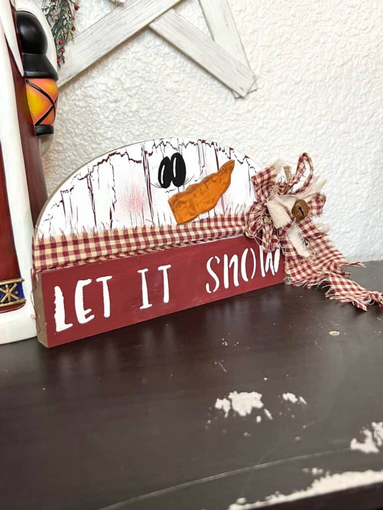 Dollar Tree half wood snowman with white paint and crackle paint underneath, cardboard carrot nose, homespun fabric scarf and bow, and the bottom portion is dark red and say let it snow.